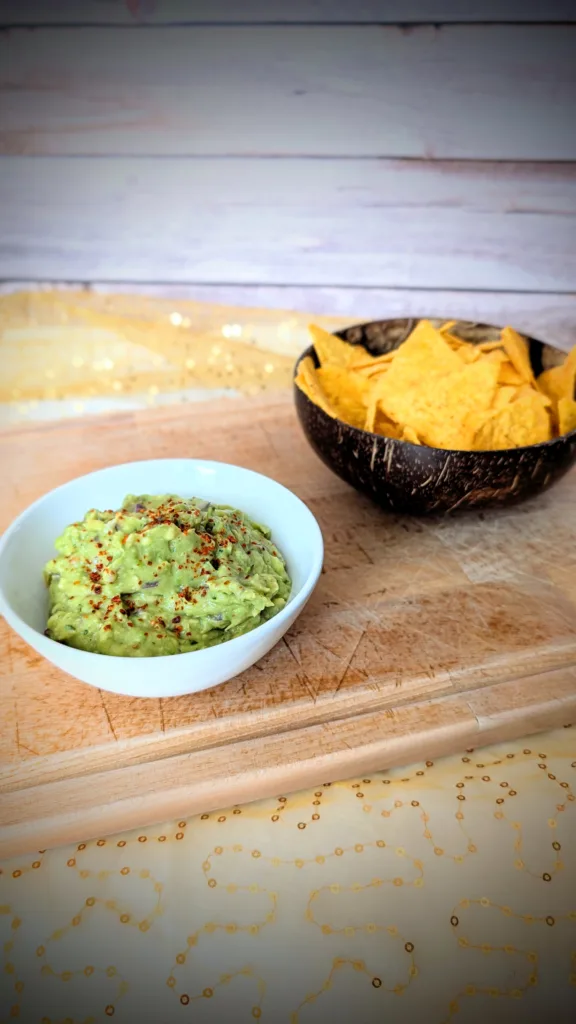 Cremige Guacamole in einer weißen Schale, mit Gewürzen bestreut und auf einem Holzbrett angerichtet. Im Hintergrund steht eine Schale mit knusprigen Tortilla-Chips – ideal zum Dippen und Servieren der selbstgemachten Guacamole.