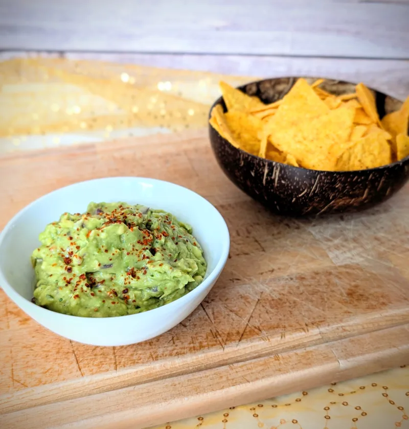 Cremige Guacamole in einer weißen Schale, mit Gewürzen bestreut und auf einem Holzbrett angerichtet. Im Hintergrund steht eine Schale mit knusprigen Tortilla-Chips – ideal zum Dippen und Servieren der selbstgemachten Guacamole.