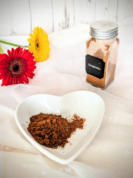 Selbstgemachtes Taco-Gewürz in einer weißen herzförmigen Schale und einem kleinen Glasbehälter vor hellem Hintergrund. Die würzige Tex-Mex-Gewürzmischung mit Paprika, Chili, Kreuzkümmel und Kakao eignet sich ideal für Tacos, Burritos und mexikanische Gerichte.