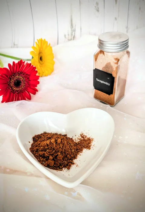 Selbstgemachtes Taco-Gewürz in einer weißen herzförmigen Schale und einem kleinen Glasbehälter vor hellem Hintergrund. Die würzige Tex-Mex-Gewürzmischung mit Paprika, Chili, Kreuzkümmel und Kakao eignet sich ideal für Tacos, Burritos und mexikanische Gerichte.