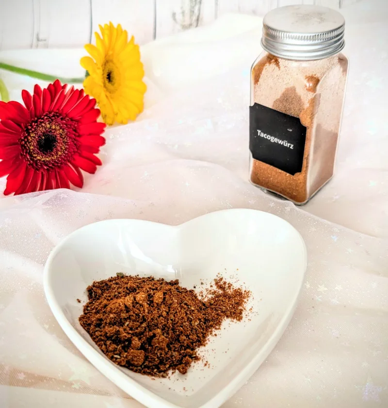 Selbstgemachtes Taco-Gewürz in einer weißen herzförmigen Schale und einem kleinen Glasbehälter vor hellem Hintergrund. Die würzige Tex-Mex-Gewürzmischung mit Paprika, Chili, Kreuzkümmel und Kakao eignet sich ideal für Tacos, Burritos und mexikanische Gerichte.