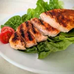 Saftige gegrillte Hähnchenbrust mit würziger Paprikakruste, auf frischen Salatblättern angerichtet und mit halbierten Cherrytomaten serviert. Die in Scheiben geschnittene Hähnchenbrust zeigt eine zarte, saftige Textur und deutliche Grillstreifen auf einem weißen Teller.