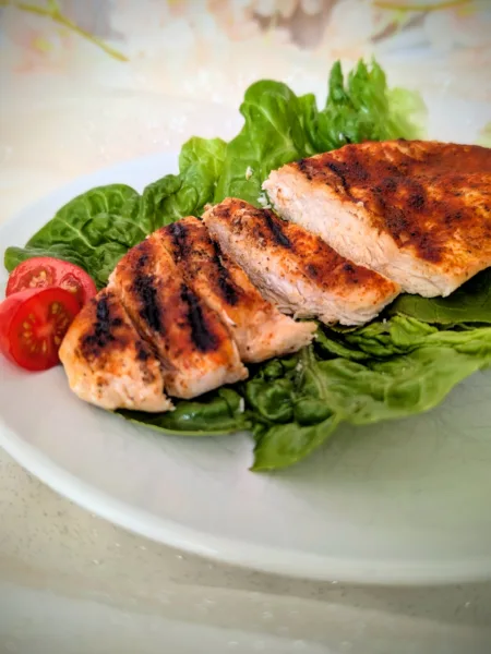 Saftige gegrillte Hähnchenbrust mit würziger Paprikakruste, auf frischen Salatblättern angerichtet und mit halbierten Cherrytomaten serviert. Die in Scheiben geschnittene Hähnchenbrust zeigt eine zarte, saftige Textur und deutliche Grillstreifen auf einem weißen Teller.