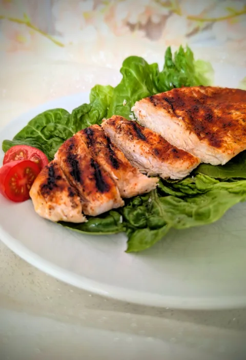 Saftige gegrillte Hähnchenbrust mit würziger Paprikakruste, auf frischen Salatblättern angerichtet und mit halbierten Cherrytomaten serviert. Die in Scheiben geschnittene Hähnchenbrust zeigt eine zarte, saftige Textur und deutliche Grillstreifen auf einem weißen Teller.