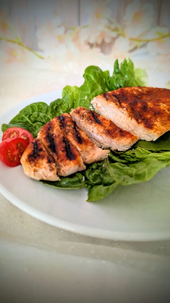 Saftige gegrillte Hähnchenbrust mit würziger Paprikakruste, auf frischen Salatblättern angerichtet und mit halbierten Cherrytomaten serviert. Die in Scheiben geschnittene Hähnchenbrust zeigt eine zarte, saftige Textur und deutliche Grillstreifen auf einem weißen Teller.