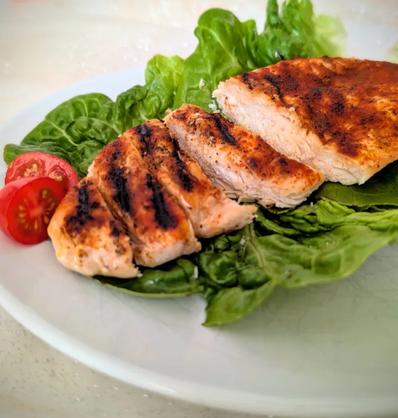 Saftige gegrillte Hähnchenbrust mit würziger Paprikakruste, auf frischen Salatblättern angerichtet und mit halbierten Cherrytomaten serviert. Die in Scheiben geschnittene Hähnchenbrust zeigt eine zarte, saftige Textur und deutliche Grillstreifen auf einem weißen Teller.